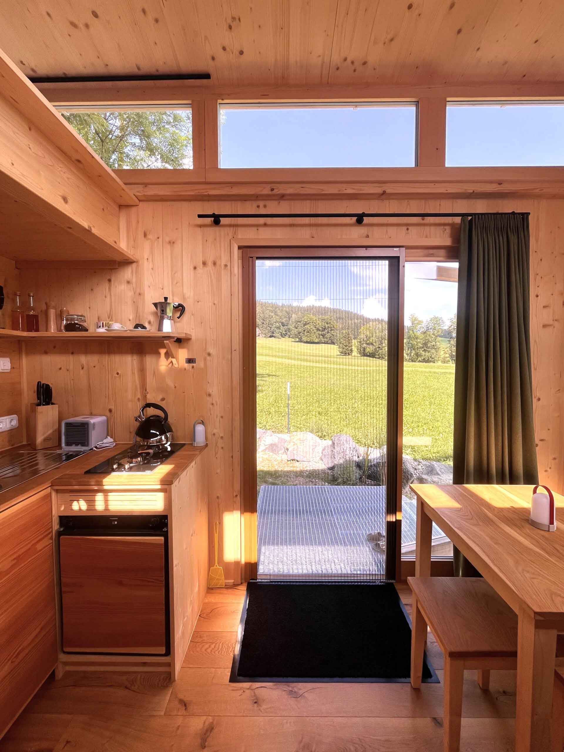 Küche und Essbereich eines Tiny Houses am Mächlerhof mit Holzverkleidung und Aussicht durch die Terrassentür auf die Allgäuer Landschaft.